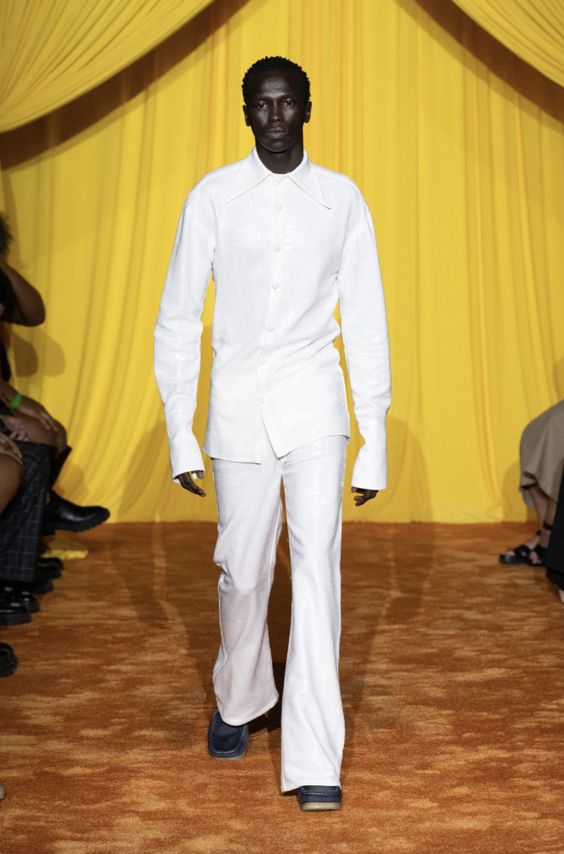 Slender male model with deep skin wearing white ensemble walking on a runway against a yellow backdrop